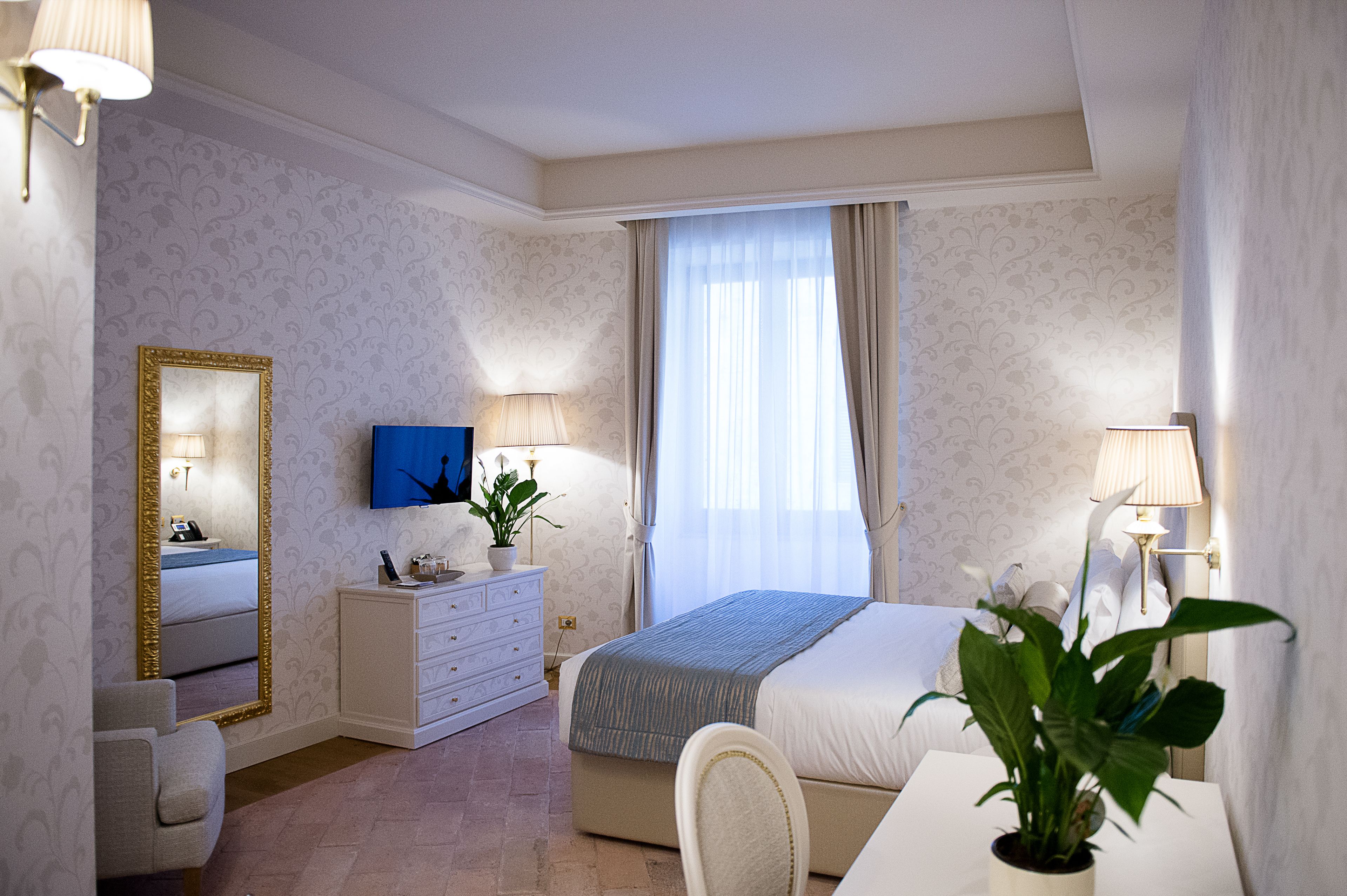 Habitación de hotel con cama, mesita de noche, silla, cómoda, espejo y planta en maceta.