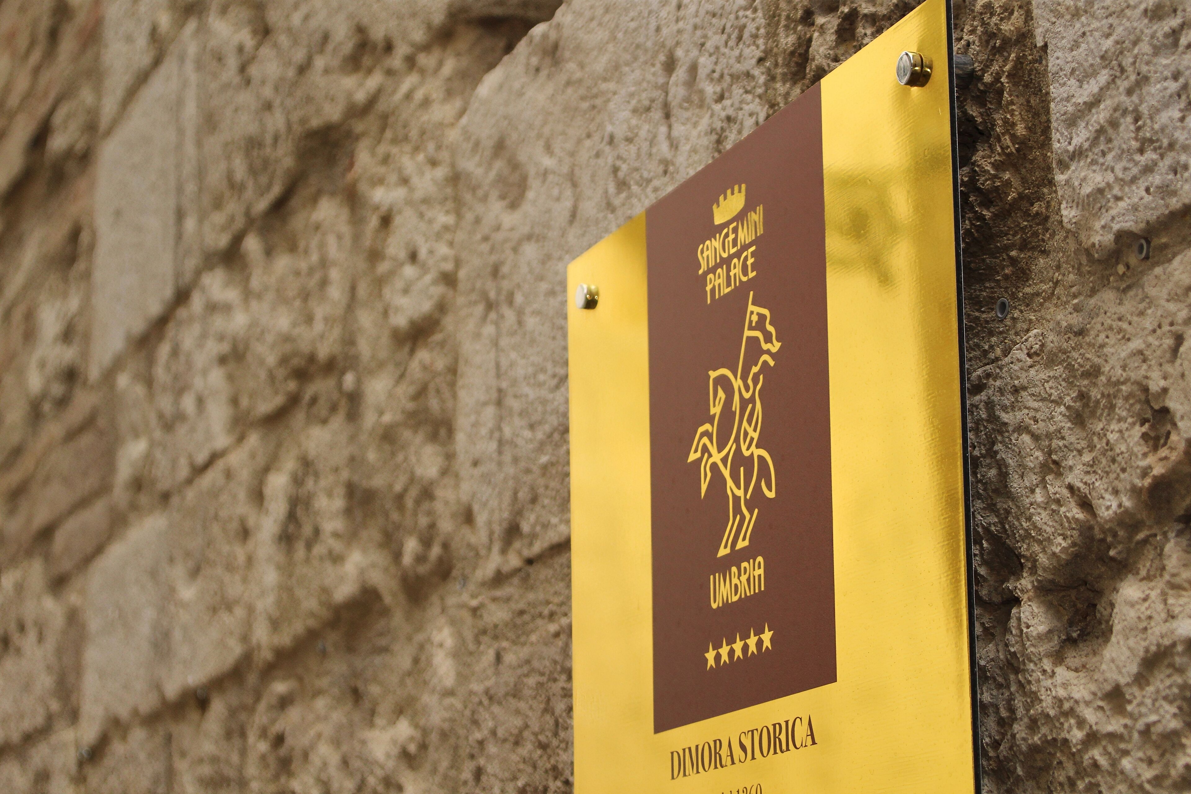 Un letrero con el texto 'SANGENMINI PALACE' y 'UMBRIA' sobre un fondo amarillo, fijado en una pared de piedra.