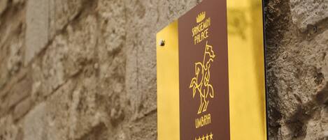 Un letrero con el texto 'SANGENMINI PALACE' y 'UMBRIA' sobre un fondo amarillo, fijado en una pared de piedra.