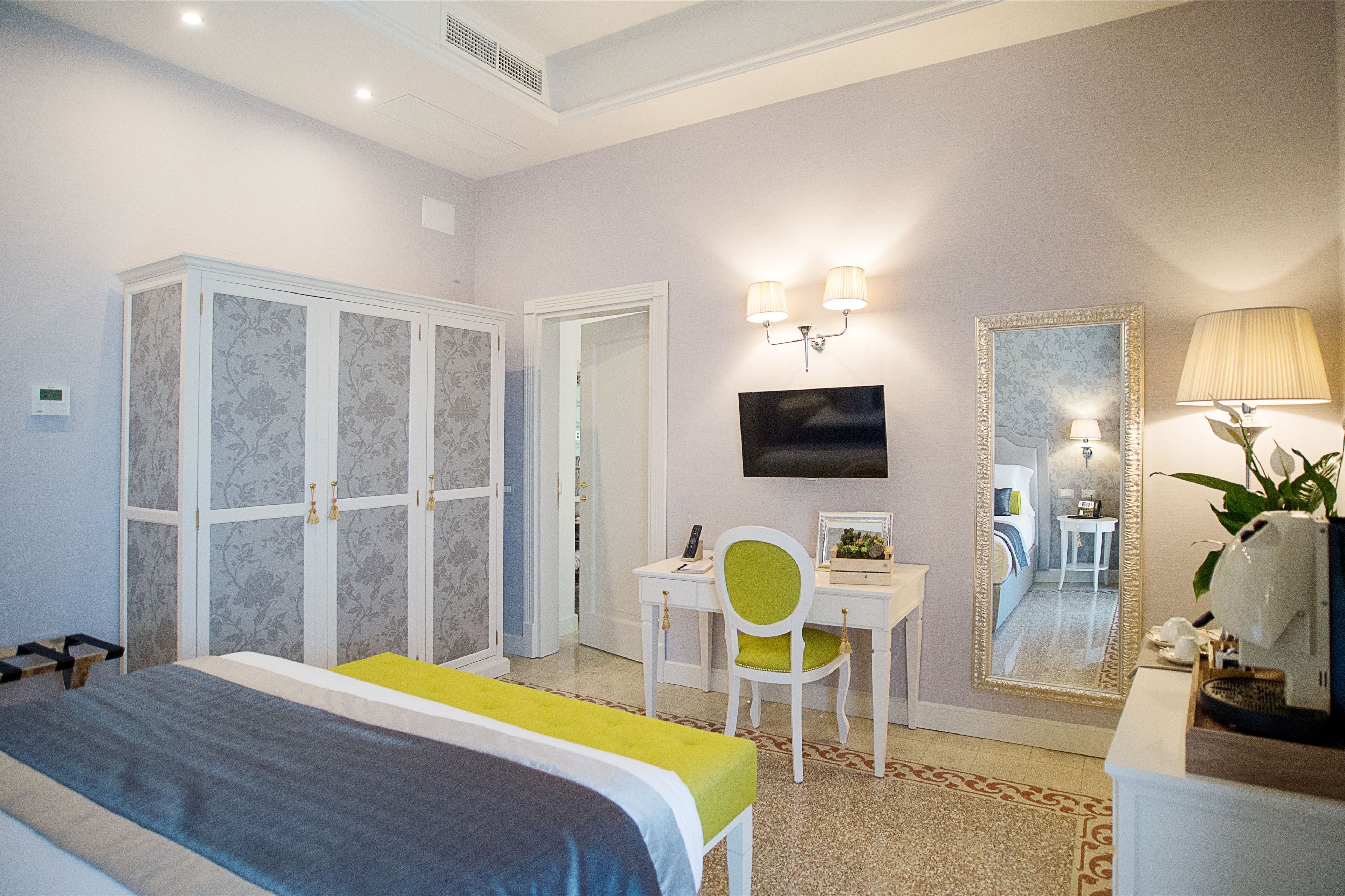 A hotel room with a bed, a desk with a chair, a television, a mirror, and a lamp.