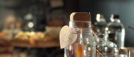Un dispensador de bebidas con tapa de corcho y etiqueta en forma de corazón, rodeado de diferentes alimentos y recipientes sobre una encimera.