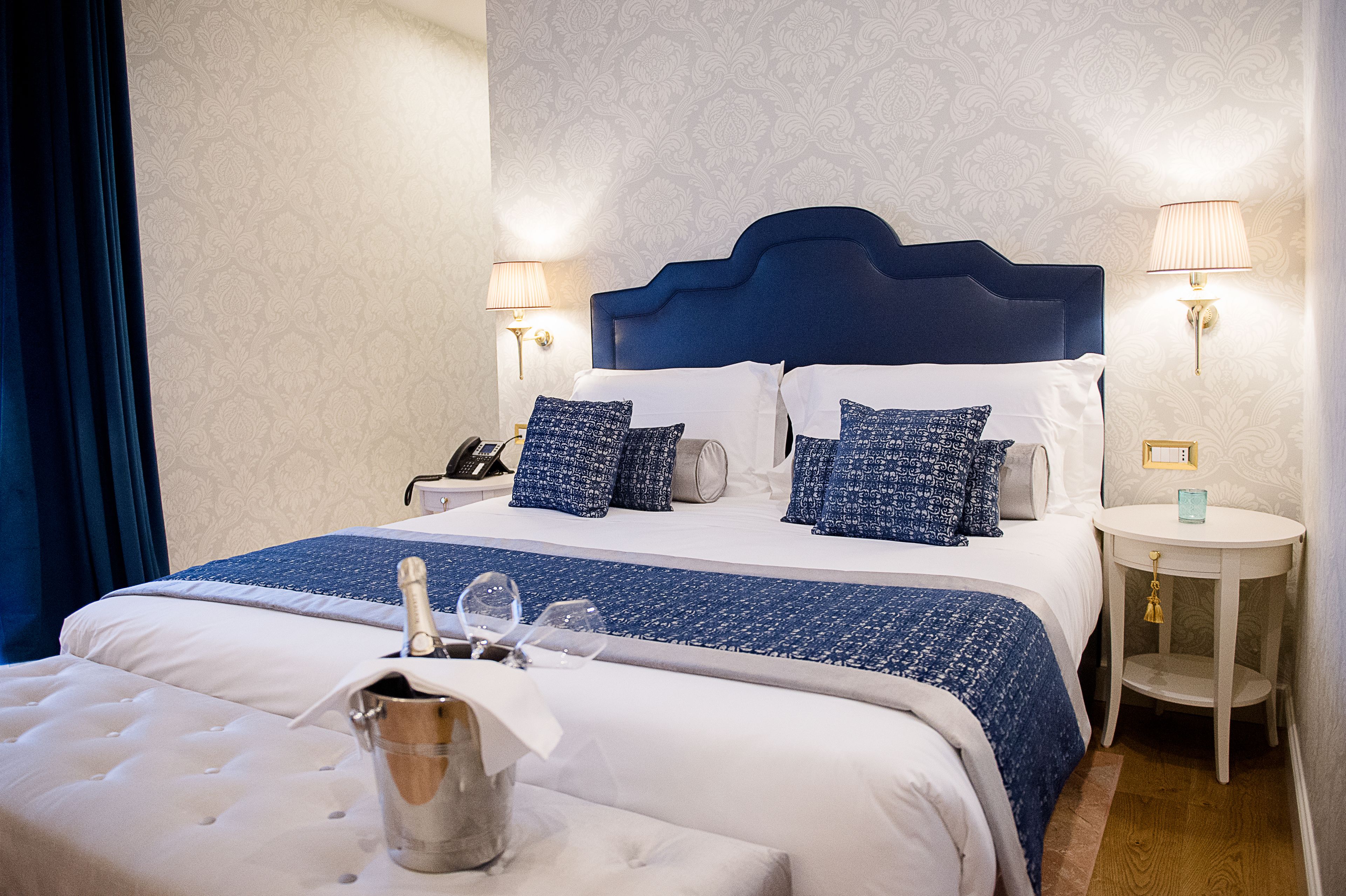 Ein ordentlich bezogenes Bett mit blauem Kopfteil, weißer Bettwäsche und gemusterten Kissen. Ein Nachttisch mit einer Lampe und einem kleinen dekorativen Gegenstand ist vorhanden.