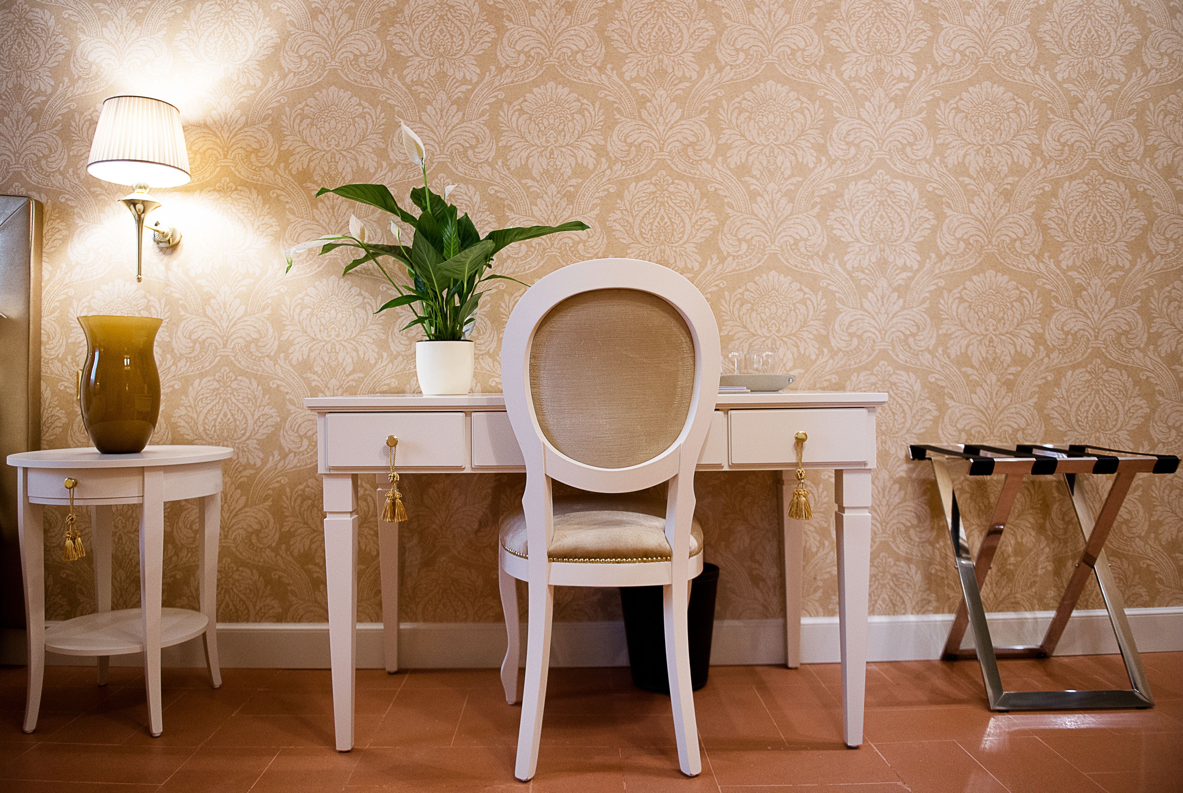 Een kamer met een wit bureau, een stoel, een kamerplant, een lamp en een bijzettafeltje met een vaas.