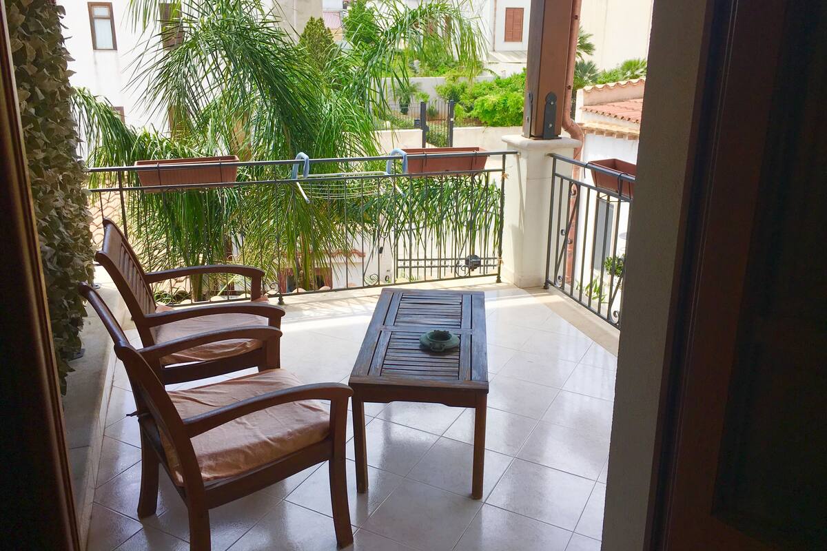 A balcony with two wooden chairs and a small table, surrounded by potted plants and a metal railing.