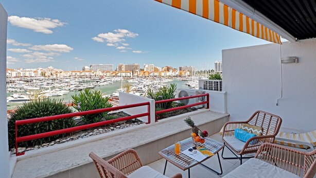 A balcony with wicker furniture, a table with a drink, and a view of a marina with boats and buildings. - Home Out Luxury Marina Vilamoura (Quarteira)
