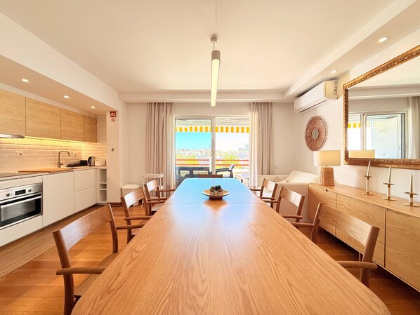 A modern kitchen with wooden cabinets, a dining area with a long wooden table, and a view of the city through large windows.