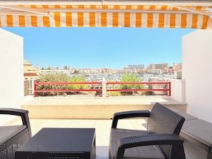 Un espace extérieur aménagé avec des meubles en osier, une rambarde rouge et une vue sur un port de plaisance et le paysage urbain.