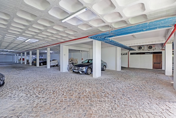 Un parking couvert, doté de plusieurs rangées de places de stationnement, de piliers blancs et d’un sol pavé.