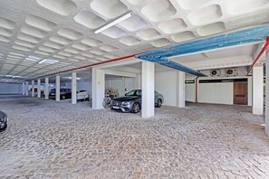 Uma garagem subterrânea coberta com várias fileiras de vagas, pilares brancos e piso de paralelepípedos.