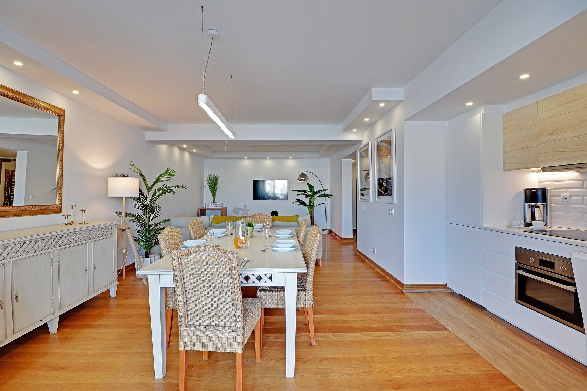 Un espace repas moderne avec une table blanche dressée pour six personnes, entourée de chaises en osier. Un grand miroir et une plante en pot se trouvent sur le côté gauche. On aperçoit la cuisine sur le côté droit, avec des armoires blanches et un four encastré.