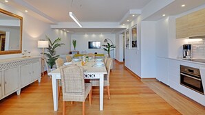 A modern dining area with a white table set for six, surrounded by wicker chairs. A large mirror and a potted plant are on the left side. The kitchen area is visible on the right with white cabinets and a built-in oven. - Home Out Luxury Marina Vilamoura (Quarteira)