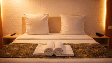 A hotel room with a bed, two pillows, a headboard, a bedside table with a phone, and a rolled towel on the bed.