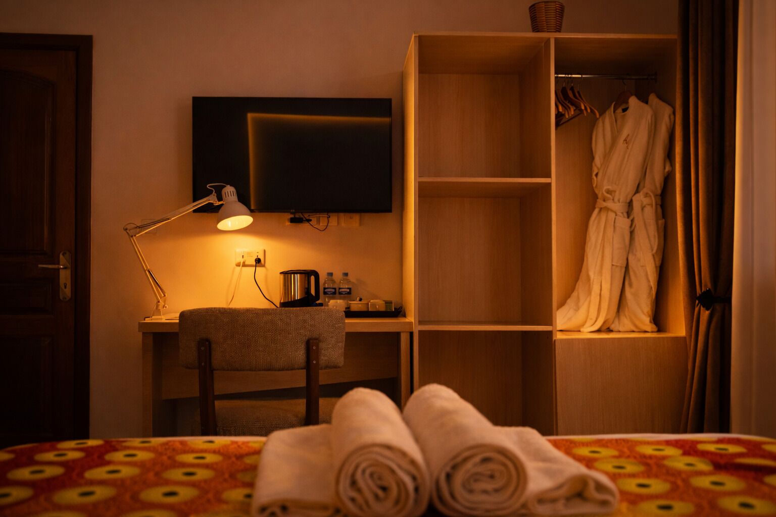 Une chambre d’hôtel avec un lit, un bureau doté d’une lampe, une télévision, une armoire et une serviette sur le lit.