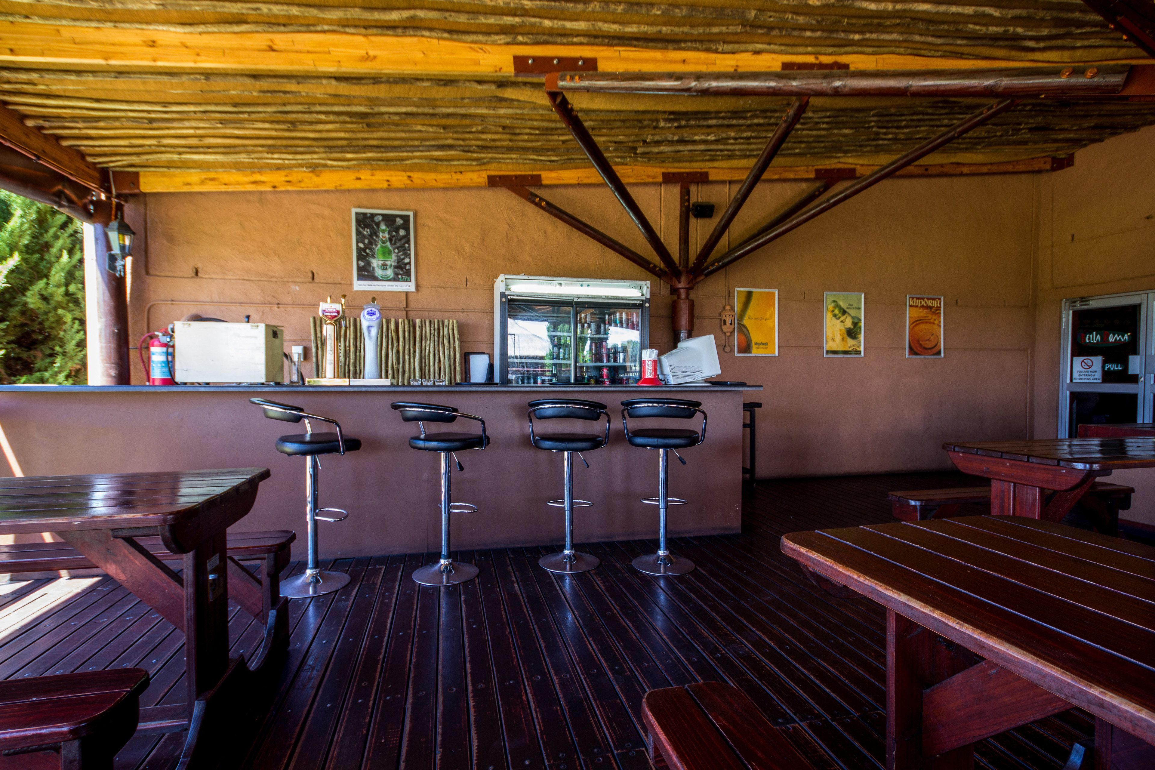 Un espace bar extérieur de style rustique, avec des tables et des tabourets en bois, un réfrigérateur rempli de boissons et une structure de plafond en bois.