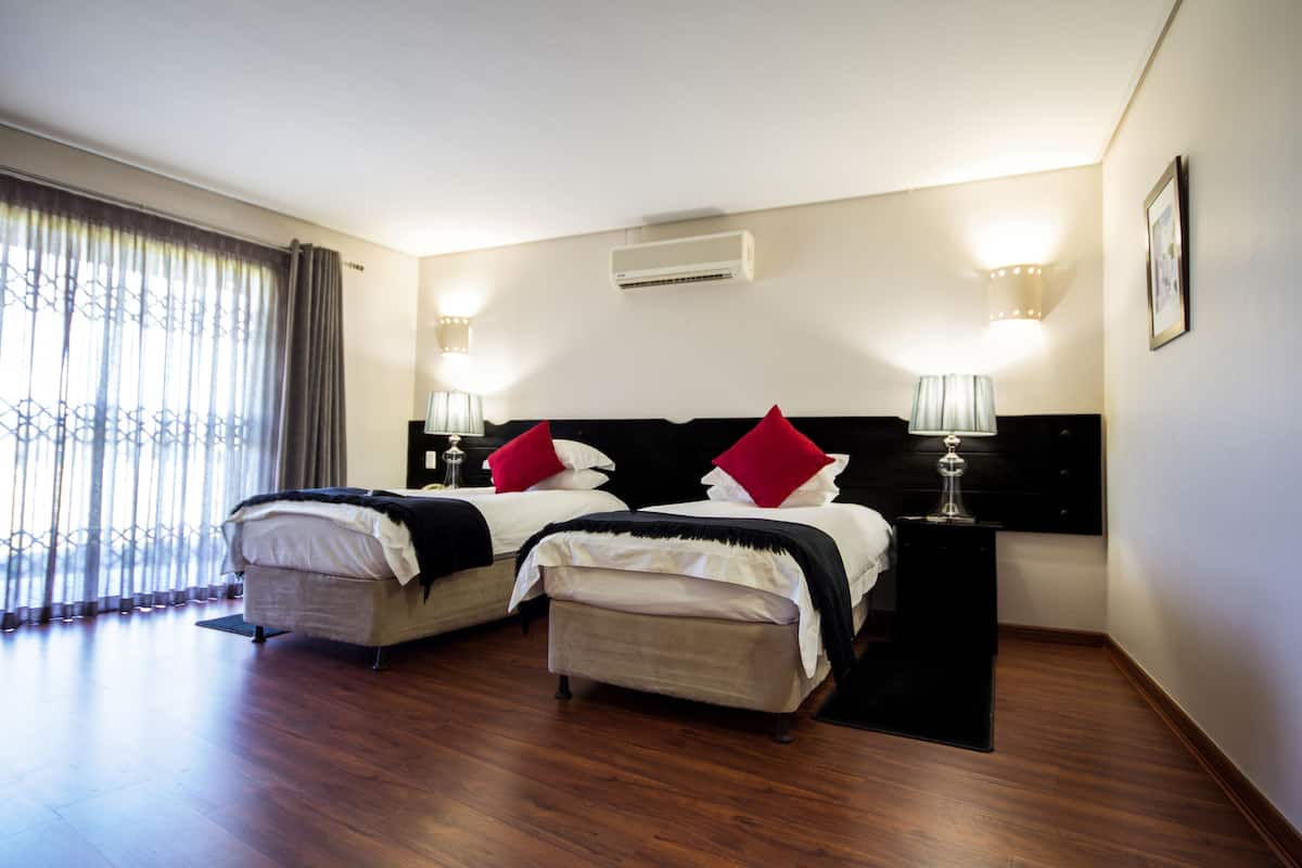 A hotel room with two beds, a nightstand, a lamp, an air conditioner, and a window with curtains.