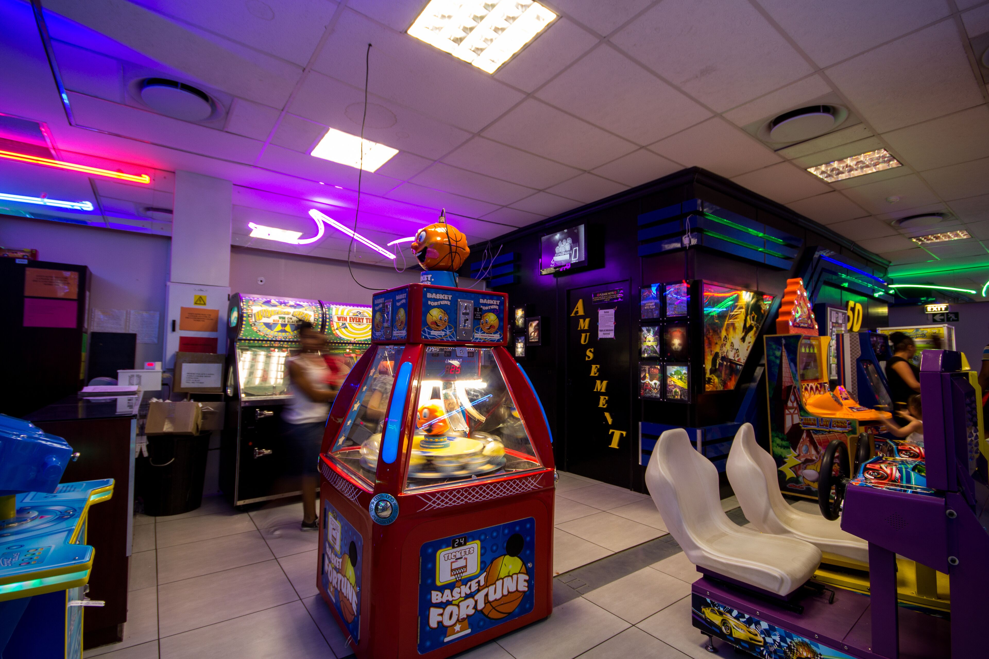 A brightly lit arcade with various gaming machines, including a basketball fortune game, and a seating area with white chairs.