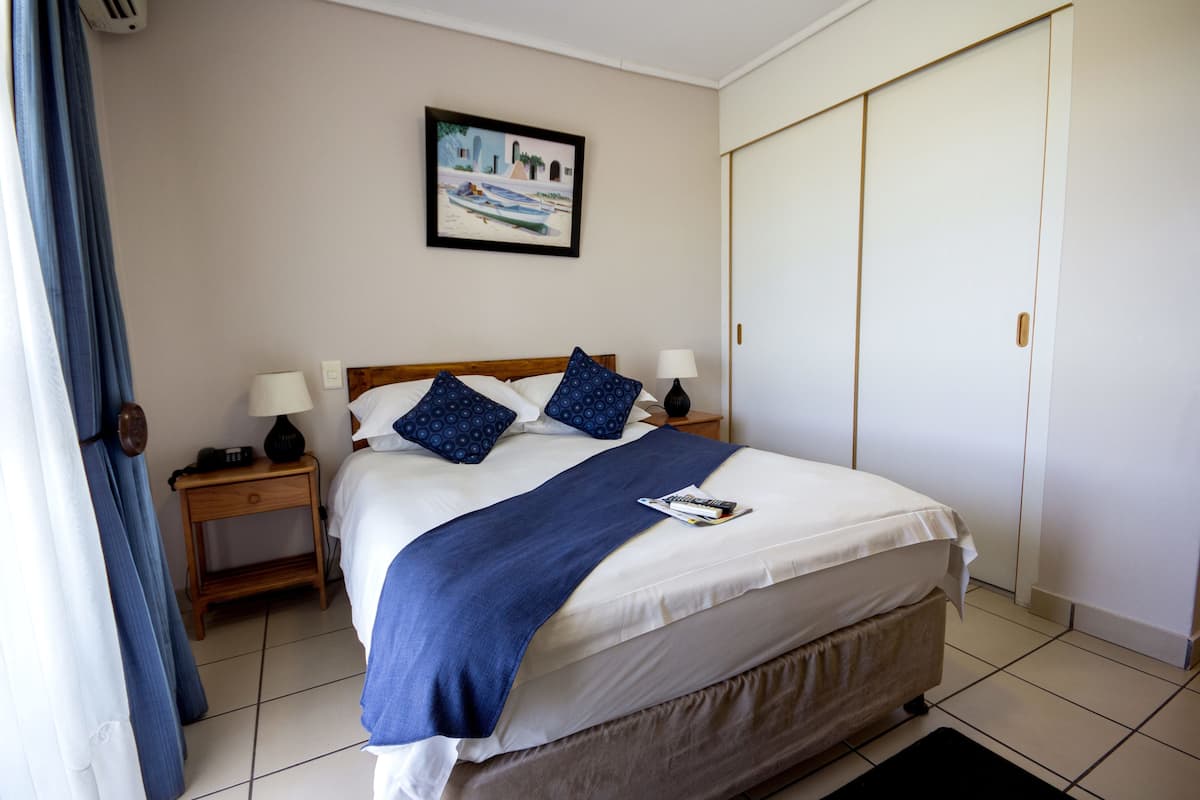 A neatly made bed with a blue blanket and pillows, a bedside table with a lamp, and a painting on the wall.