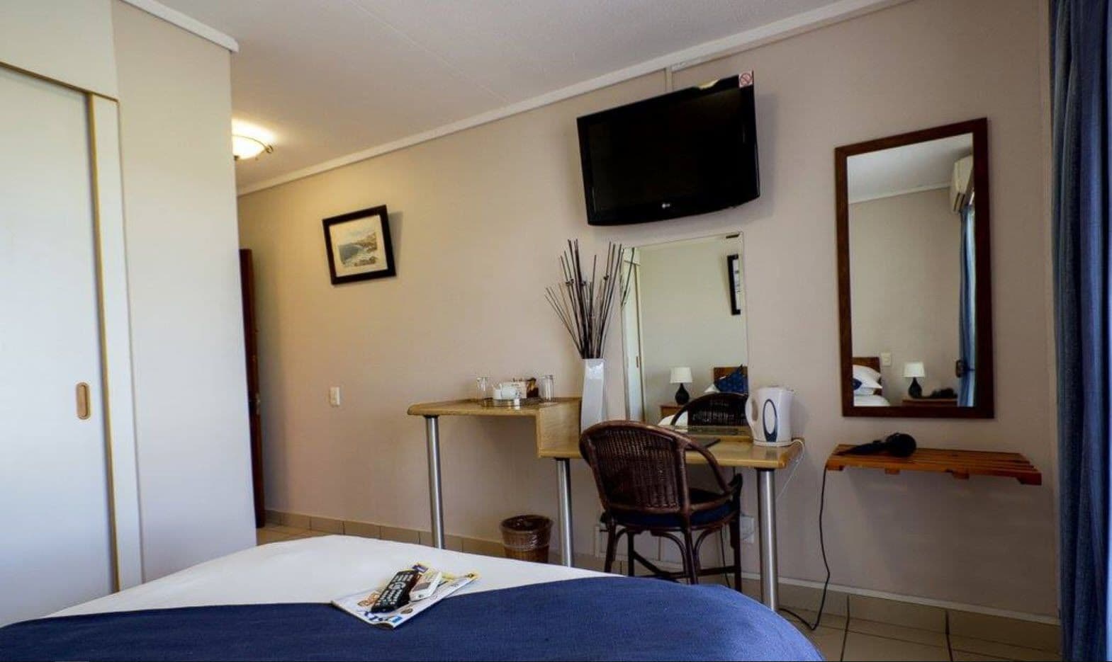 Une chambre d’hôtel comprenant un lit, un bureau avec une chaise, une télévision, un miroir et une petite table sur laquelle se trouvent un vase et une lampe.