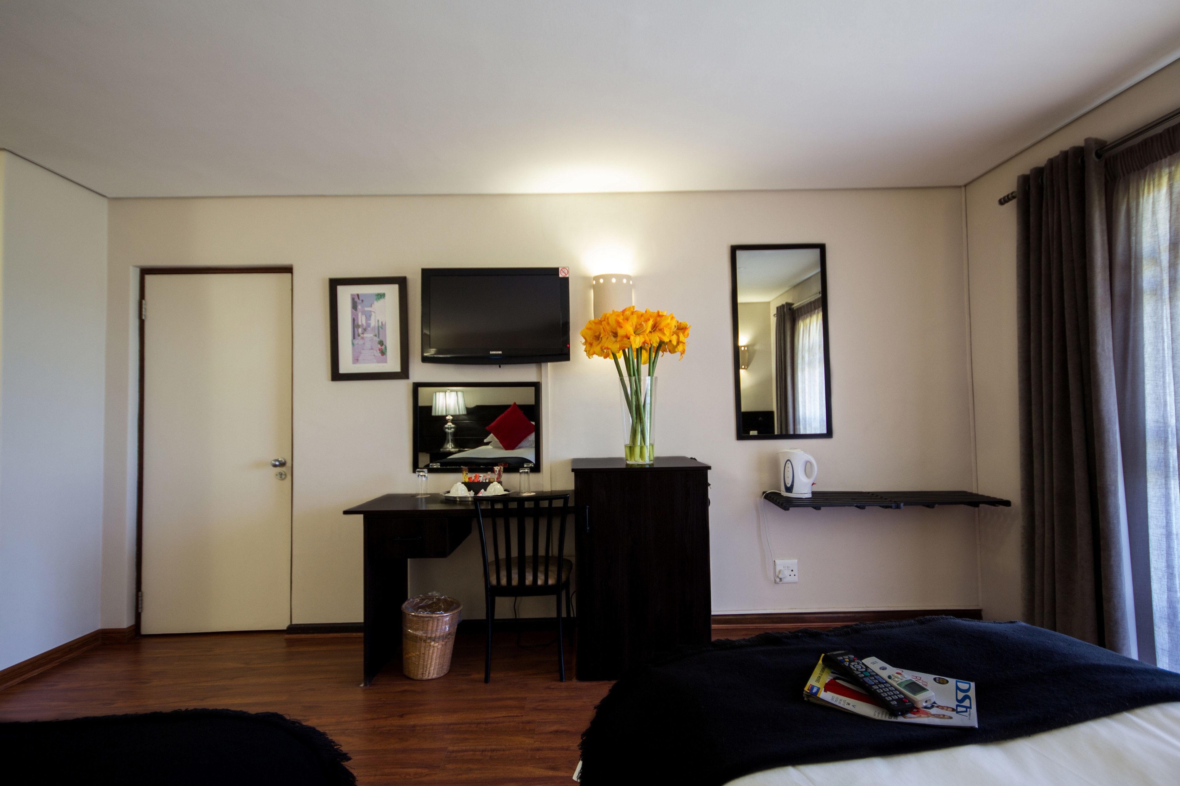 Ein Hotelzimmer mit Bett, Schreibtisch und Stuhl, Fernseher, Blumenvase, Spiegel und einem Fenster mit Vorhängen.