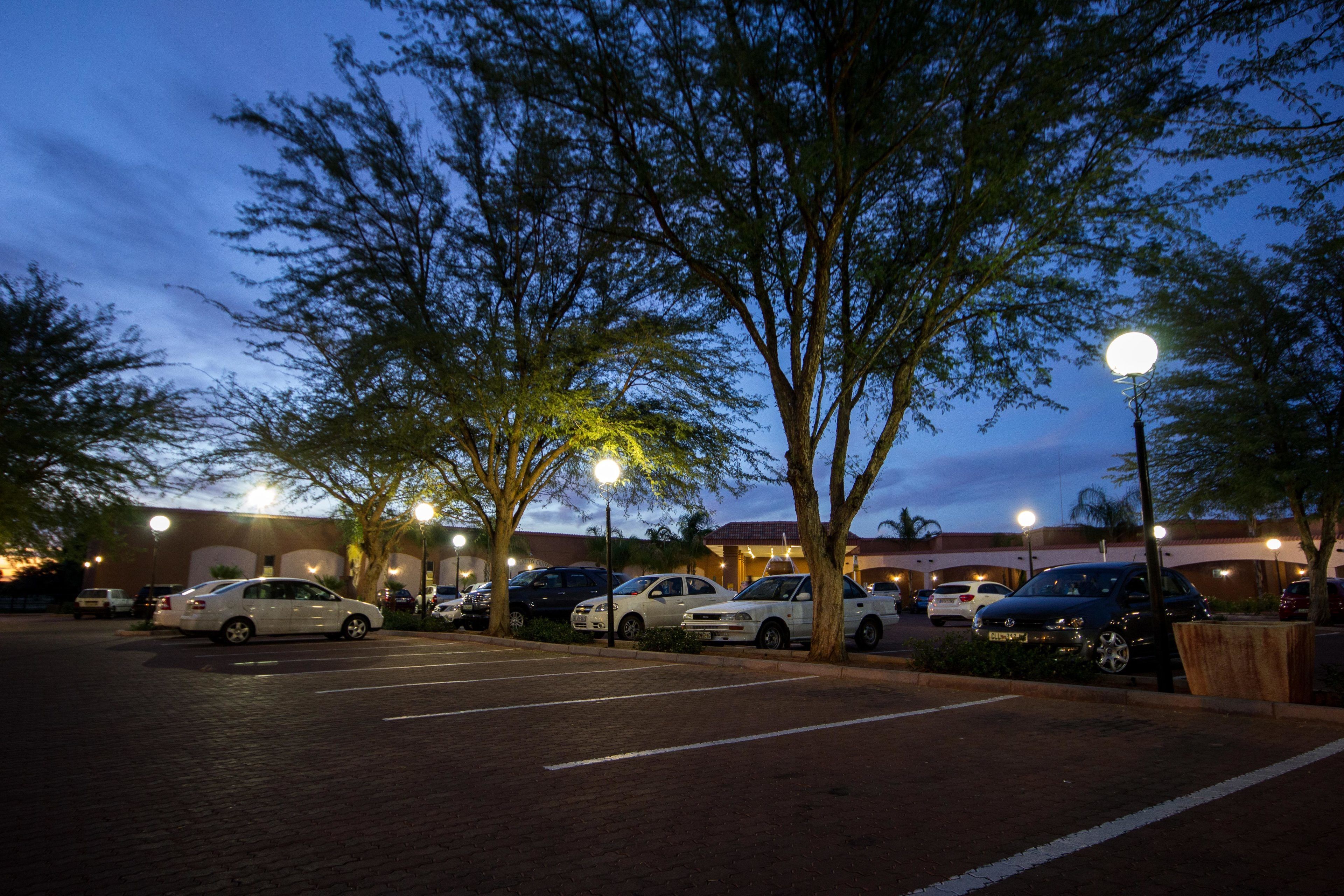 A parking lot with several cars parked, trees, and a building with lights in the background.