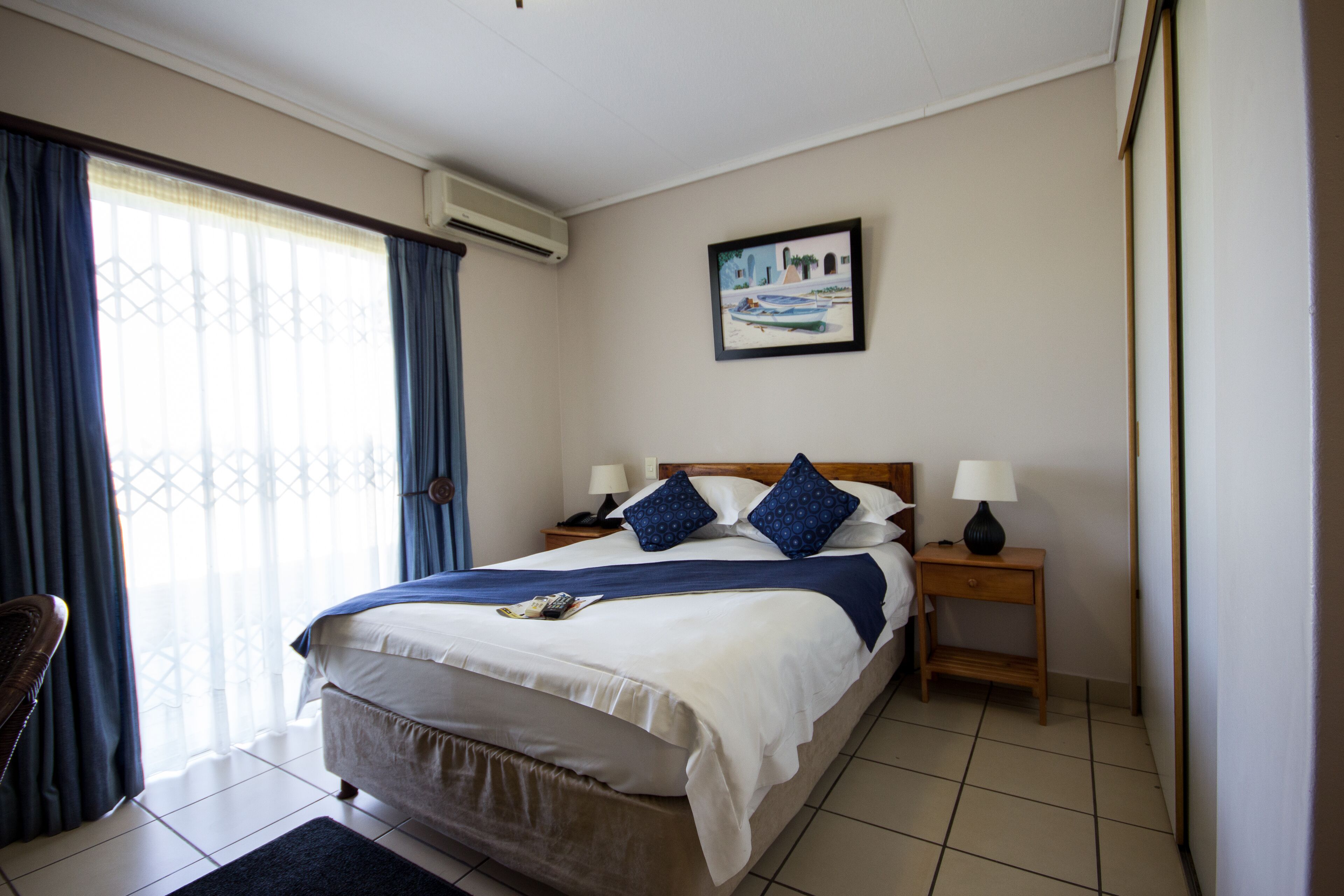 A hotel room with a bed, two bedside tables with lamps, a window with curtains, a painting on the wall, and a chair.