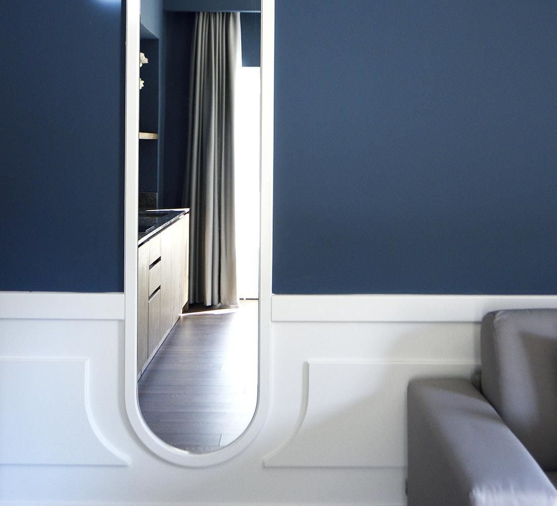 A room with a large mirror reflecting a hallway with a window, a grey sofa, and a white wall with a decorative molding.