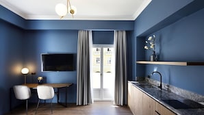 A modern kitchen with a dark blue wall, a wooden dining table, two chairs, a TV mounted above the table, and a window with curtains. - Roomore Apartments (Thessaloniki)