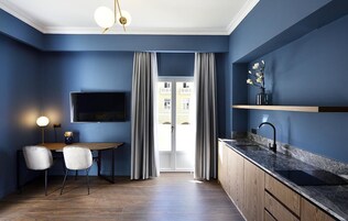 A modern kitchen with a dark blue wall, a wooden dining table, two chairs, a TV mounted above the table, and a window with curtains.