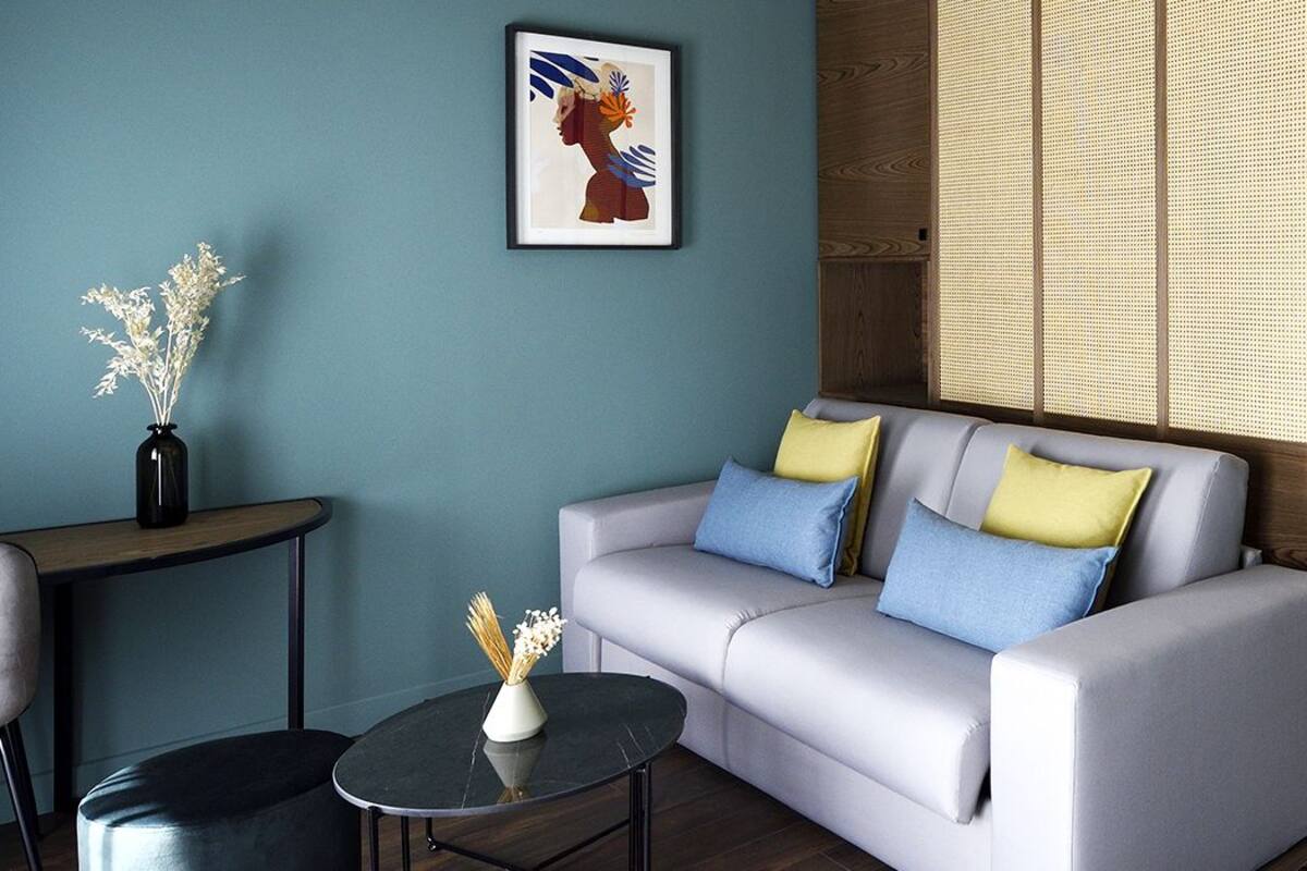 Un salon moderne avec un canapé gris, une petite table ronde et un fauteuil. Une œuvre d’art encadrée est accrochée au mur, et une fenêtre avec des stores est visible.