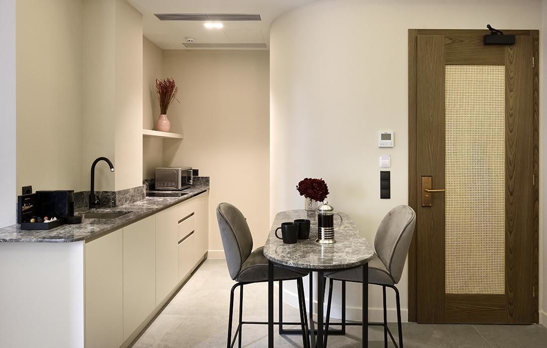 A compact kitchen with a marble countertop, a sink, a microwave, and a small shelf with a vase. There is a round table with two chairs and a vase with red flowers. A wooden door with a small window is visible.