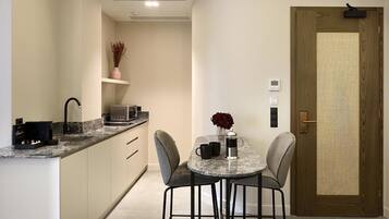 A compact kitchen with a marble countertop, a sink, a microwave, and a small shelf with a vase. There is a round table with two chairs and a vase with red flowers. A wooden door with a small window is visible.