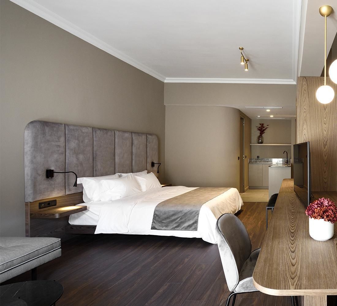 A modern hotel room with a large bed, a desk, a chair, a TV, and a small kitchen area.