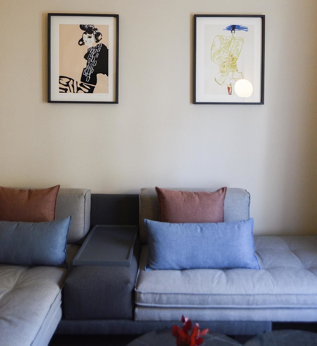 Ein modernes Wohnzimmer mit einer grauen Sofagarnitur, zwei gerahmten Kunstwerken an der Wand und einem kleinen Tisch mit einem roten Dekorationsgegenstand.