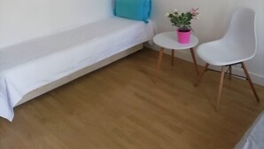 Una habitación pequeña y ordenada con una cama individual, una mesita con una planta, una silla blanca y una lámpara fijada a la pared.