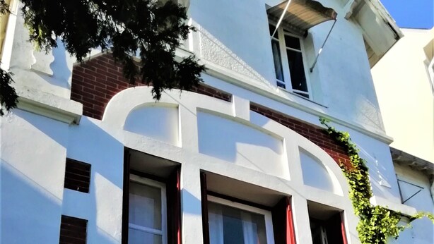 Un edificio de varios pisos con fachada blanca, múltiples ventanas con cortinas rojas y un balcón con macetas.