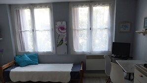 Un dormitorio con cama, escritorio y silla. Hay dos ventanas con cortinas, un radiador y un cuadro en la pared.
