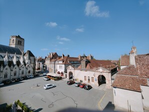 Une place historique, où se côtoient l’architecture gothique et l’architecture traditionnelle, avec notamment une église imposante dotée d’un clocher. On y voit plusieurs voitures garées et un ciel bleu dégagé au-dessus.