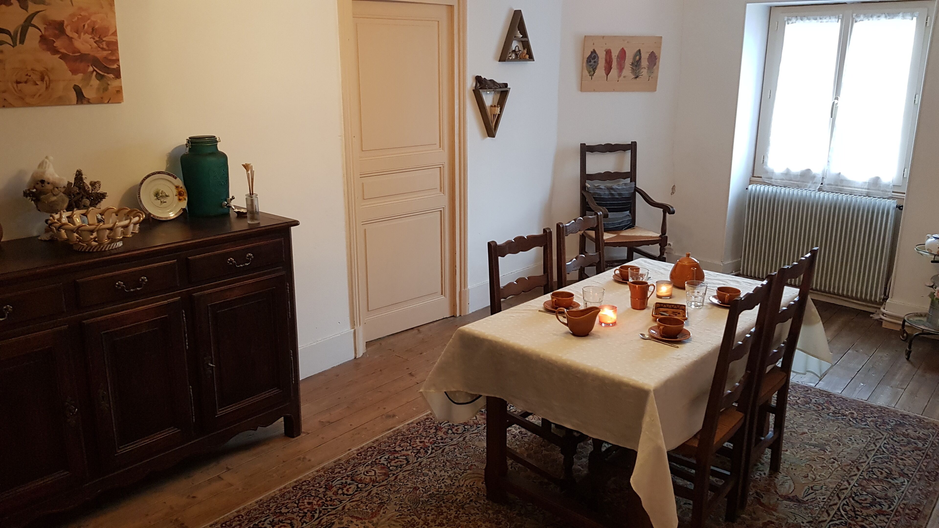 Un coin repas avec une table dressée pour quatre personnes, entourée de chaises en bois. Un tapis à motifs recouvre le sol, et un radiateur est visible au fond.