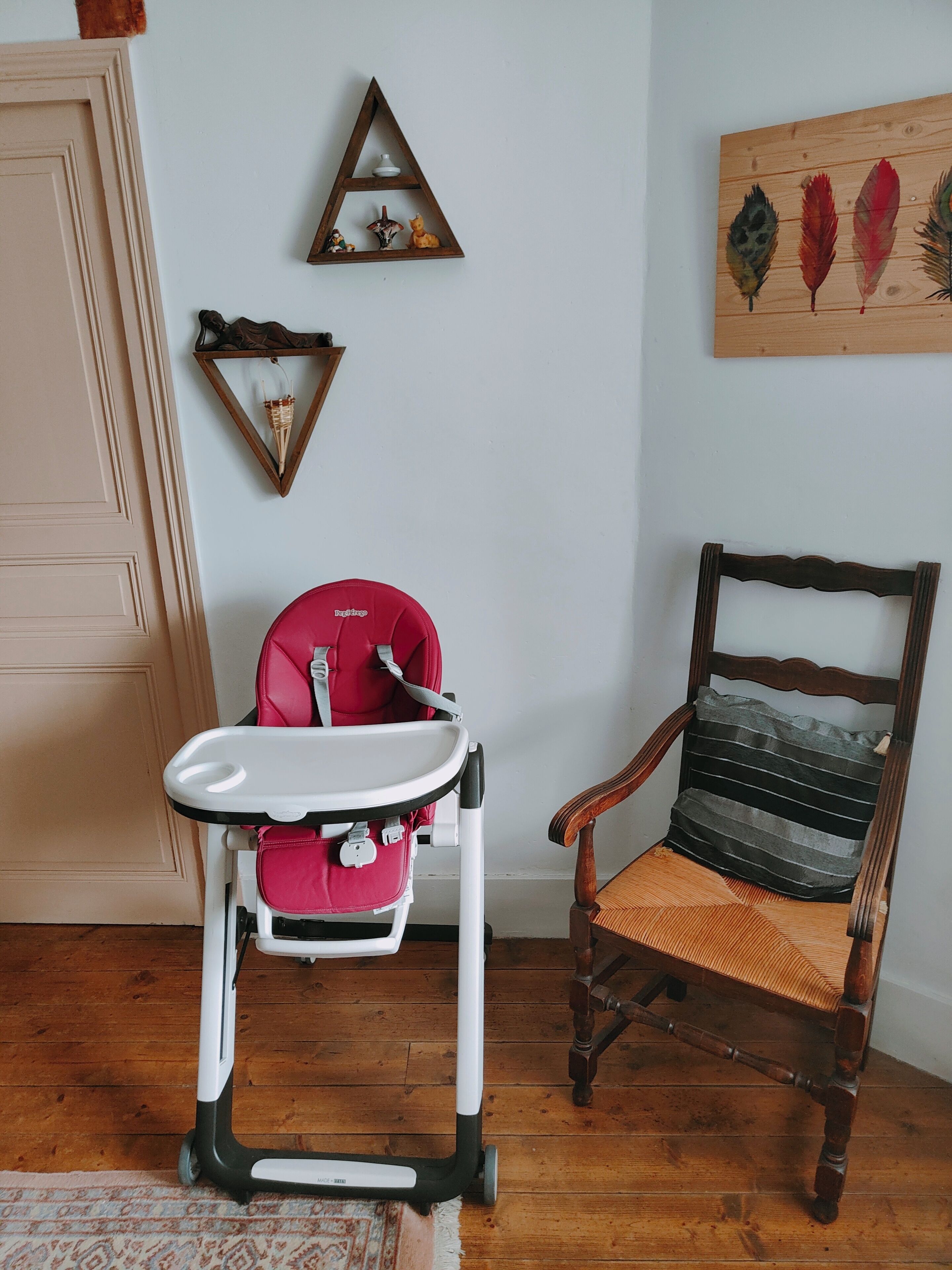 Een kamer met een kinderstoel, een houten stoel met kussen, een wandplank met decoratieve objecten, en een houten vloer.