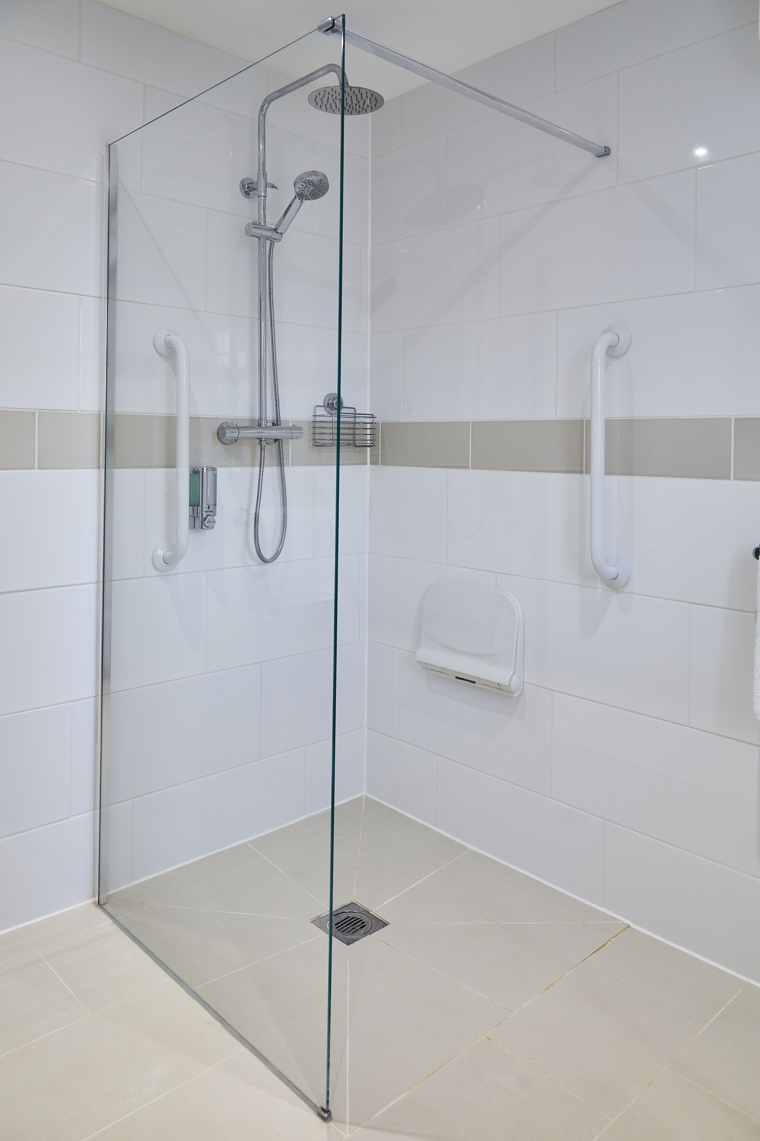 A glass-enclosed shower stall with a handheld showerhead, a wall-mounted towel dispenser, and a floor drain.