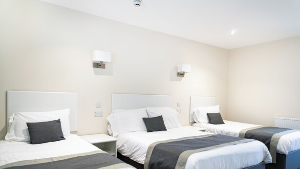 A hotel room with three beds, each with a white headboard and grey bedding. There are two wall-mounted light fixtures and a ceiling light. - Appleby Inn Hotel (Swadlincote)