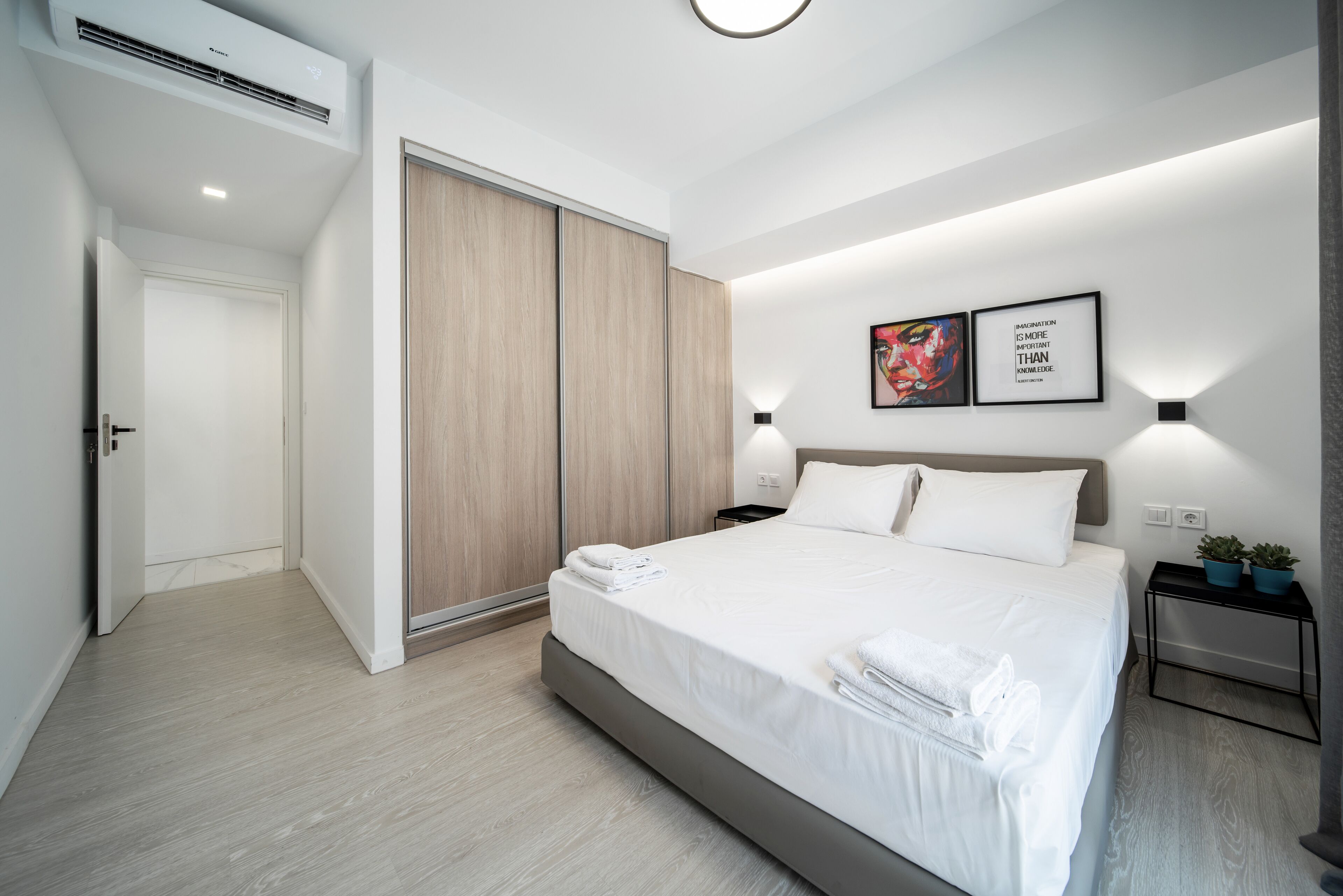 A modern hotel room with a large bed, a wardrobe, and a bedside table with two potted plants.