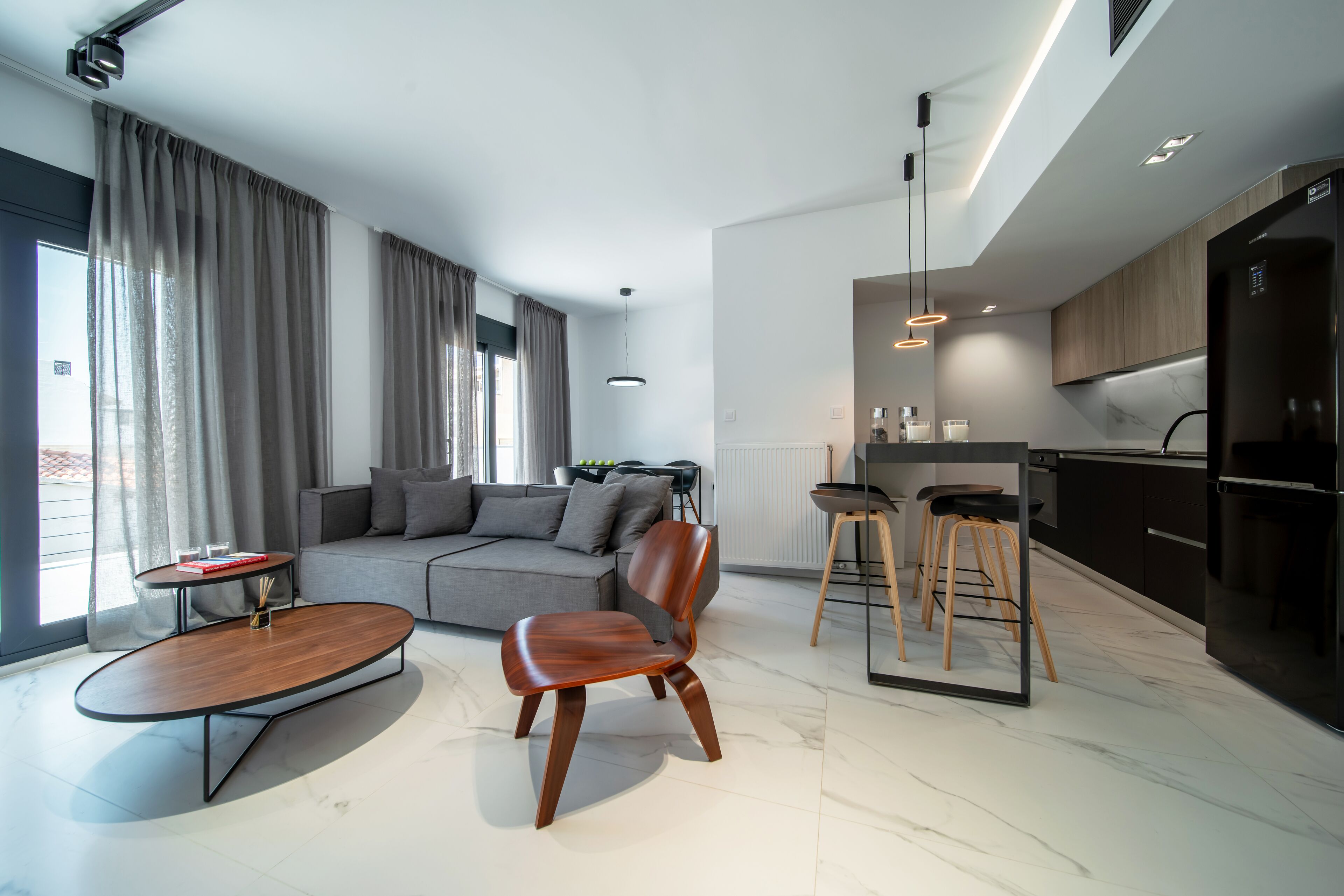A modern living room with a sofa, a coffee table, and a chair. There is a kitchen area with a refrigerator and bar stools.