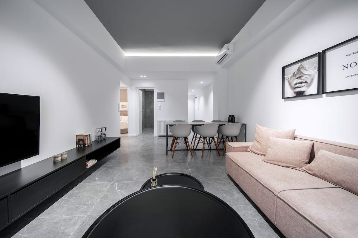Uma sala de estar moderna com uma TV de tela plana, uma mesa de centro preta, um sofá bege e uma área de jantar com mesa e cadeiras.