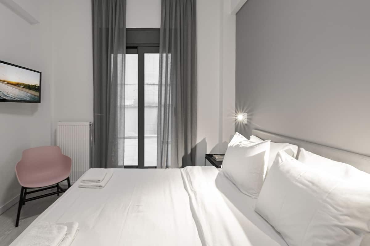 Quarto de hotel moderno com uma cama grande, uma cadeira, uma mesa de cabeceira e vista para o exterior através de uma janela com cortinas.