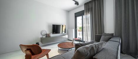 Una sala de estar moderna con un sofá grande, una silla de madera, un televisor sobre un mueble blanco y una ventana con cortinas.