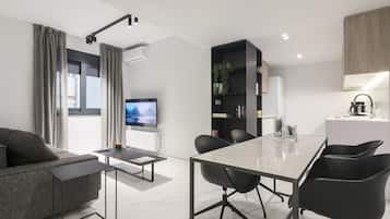 A modern living room with a sofa, a coffee table, and a TV. There is a dining area with a table and chairs. A bookshelf and a kitchen area are also visible.