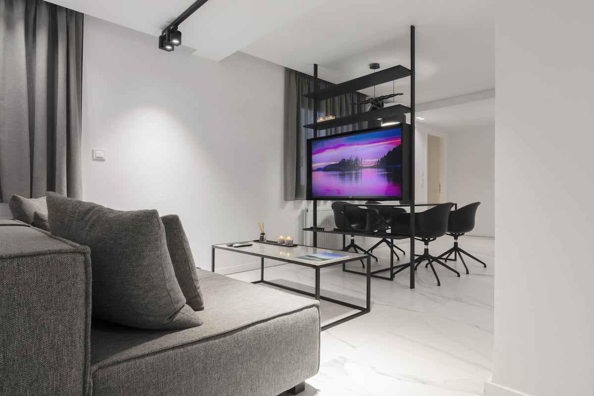Uma sala de estar moderna com um sofá cinza, uma mesa de centro, uma TV presa a um suporte preto, e uma área de jantar com cadeiras e mesa pretas.