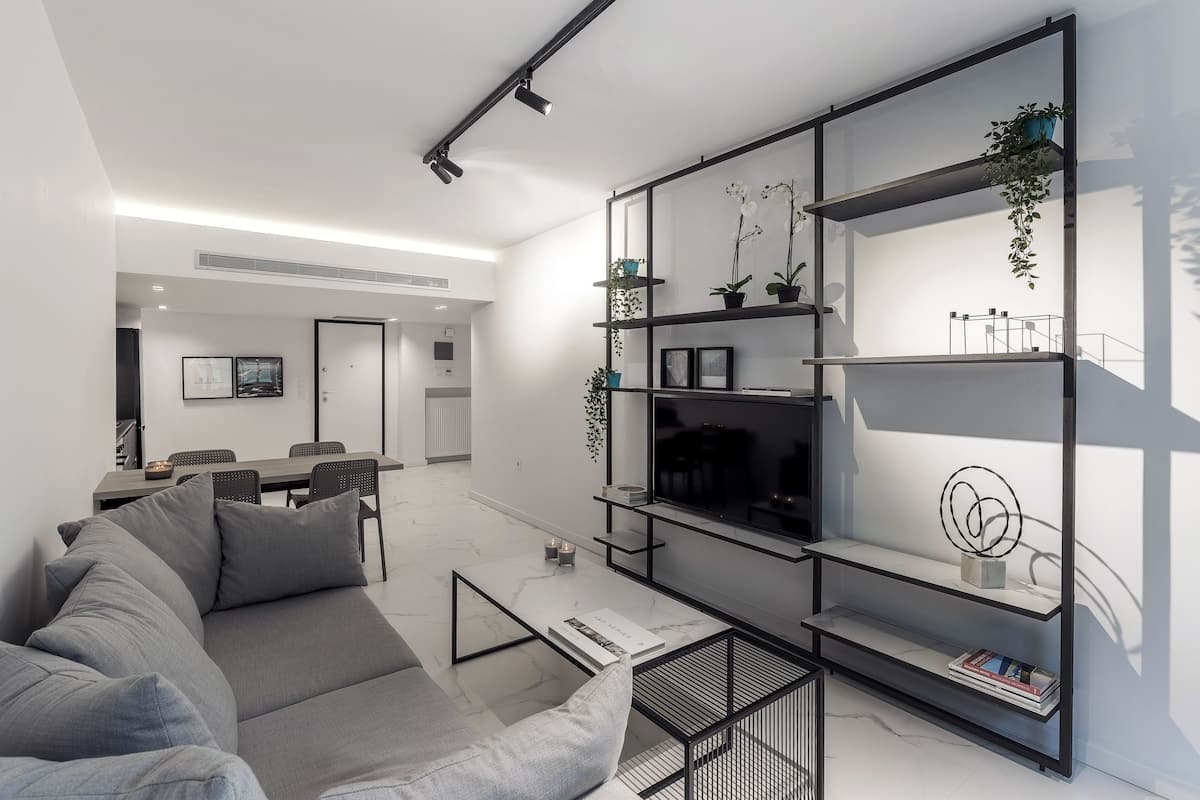 Uma sala de estar moderna com um sofá cinzento, uma mesa de centro e um suporte de TV preto de metal com prateleiras. Ao fundo, há uma área de jantar com uma mesa e cadeiras.