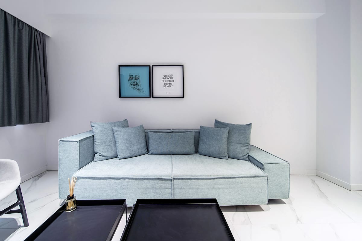 Uma sala de estar moderna com um sofá azul claro, duas frases emolduradas na parede e uma mesa de centro preta em primeiro plano.